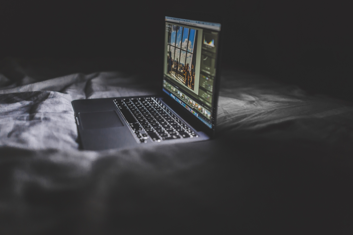 Image: A Macbook Pro sits on a bed in a darkened room. The screen shines in the dark.