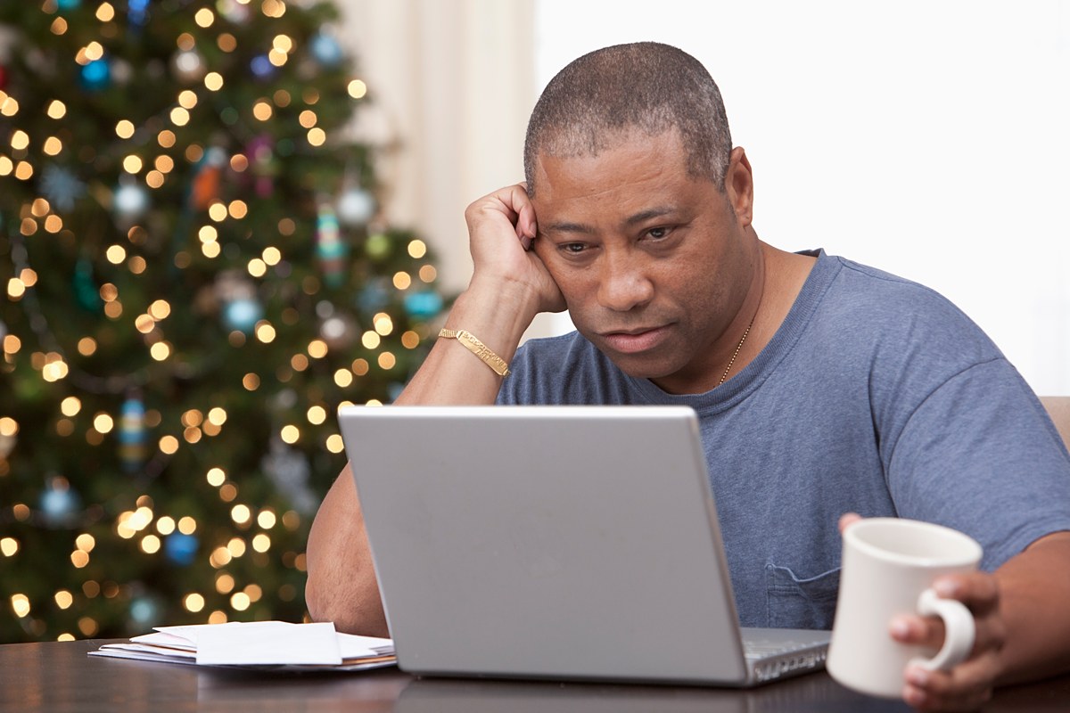 A sleepy-looking man with a mug of coffee works on a laptop with a Christmas tree in the background.