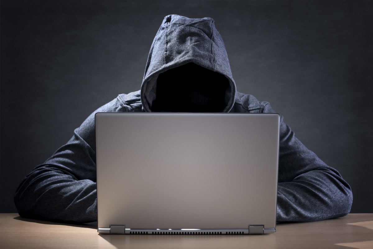 A hacker, wearing a hooded jacket and face obscured, sits in front of a laptop.'
