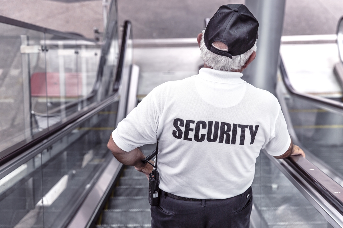 A security guard travels down an elevator. From behind, we see the back of his shirt with 'Security' in big bold letters.